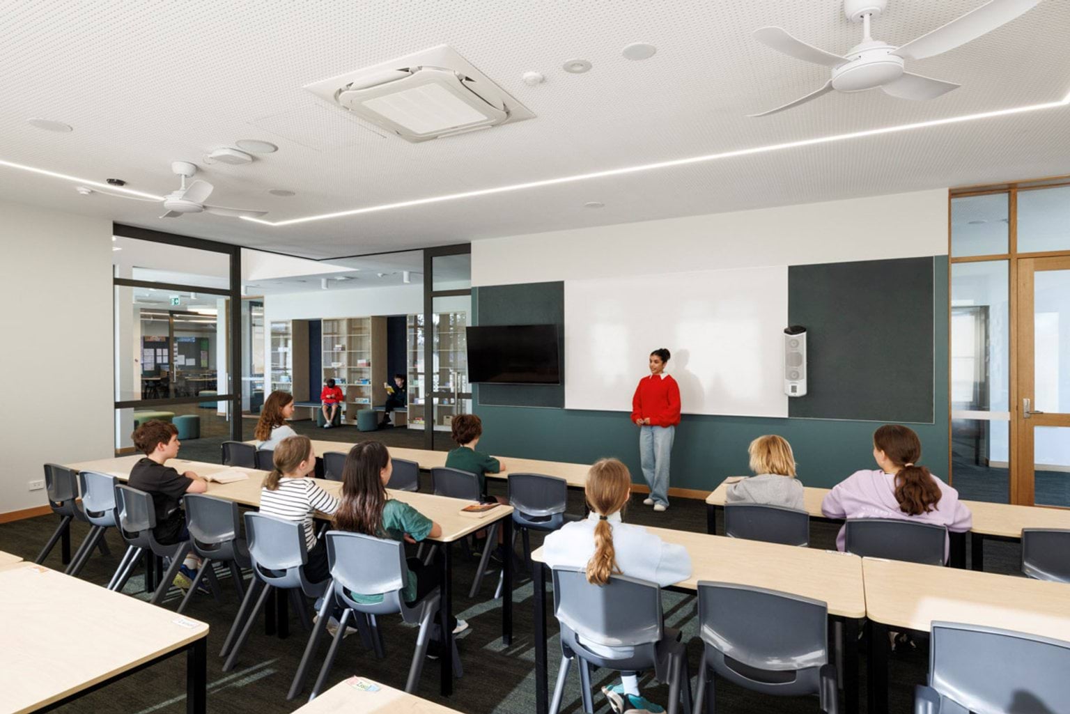 A bright classroom with rows of desks facing a whiteboard and wall-mounted screen. A teacher stands at the front while students sit at tables.
