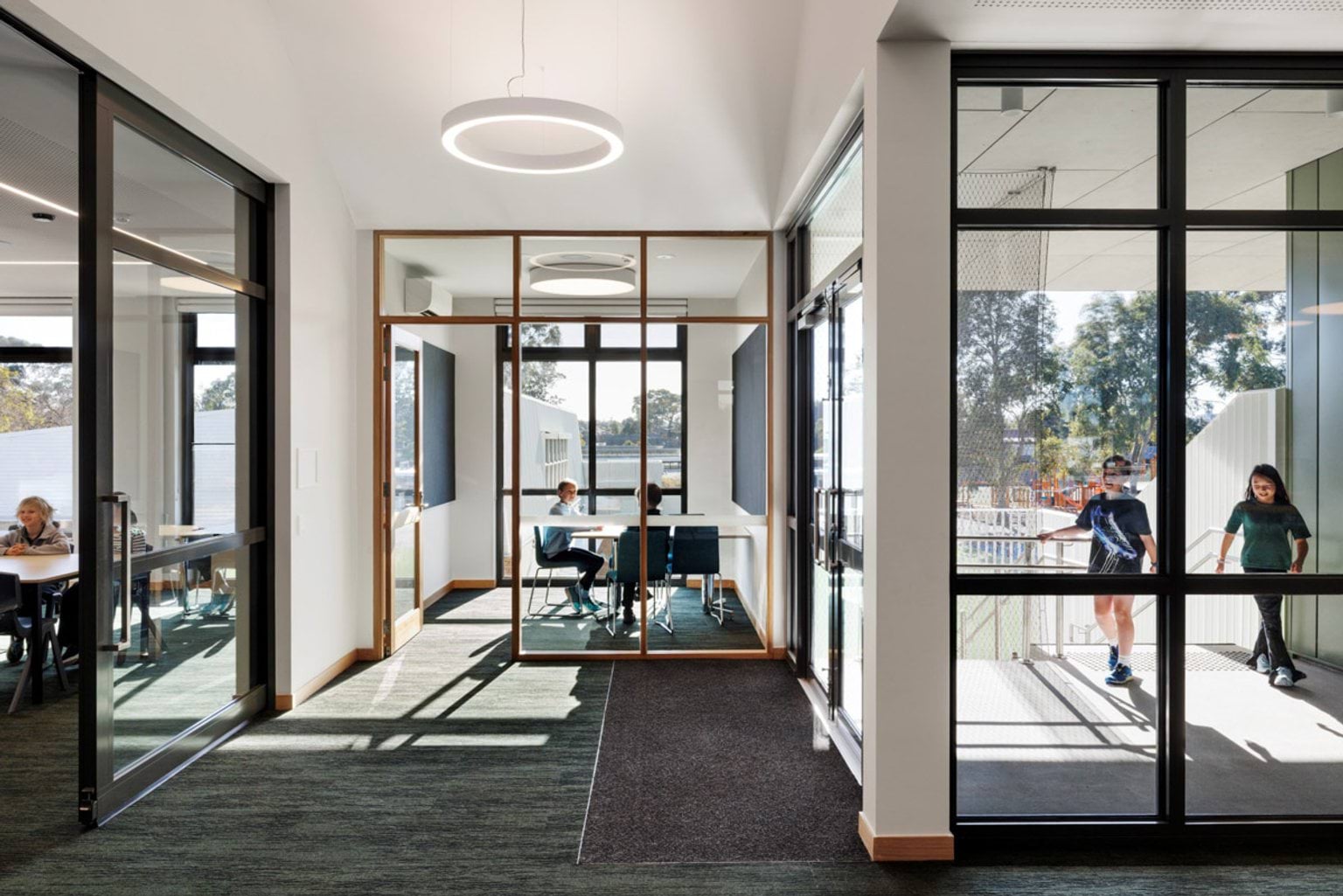 A glass-walled meeting room with a round light fixture, adjacent to a corridor with large windows and carpeted flooring.