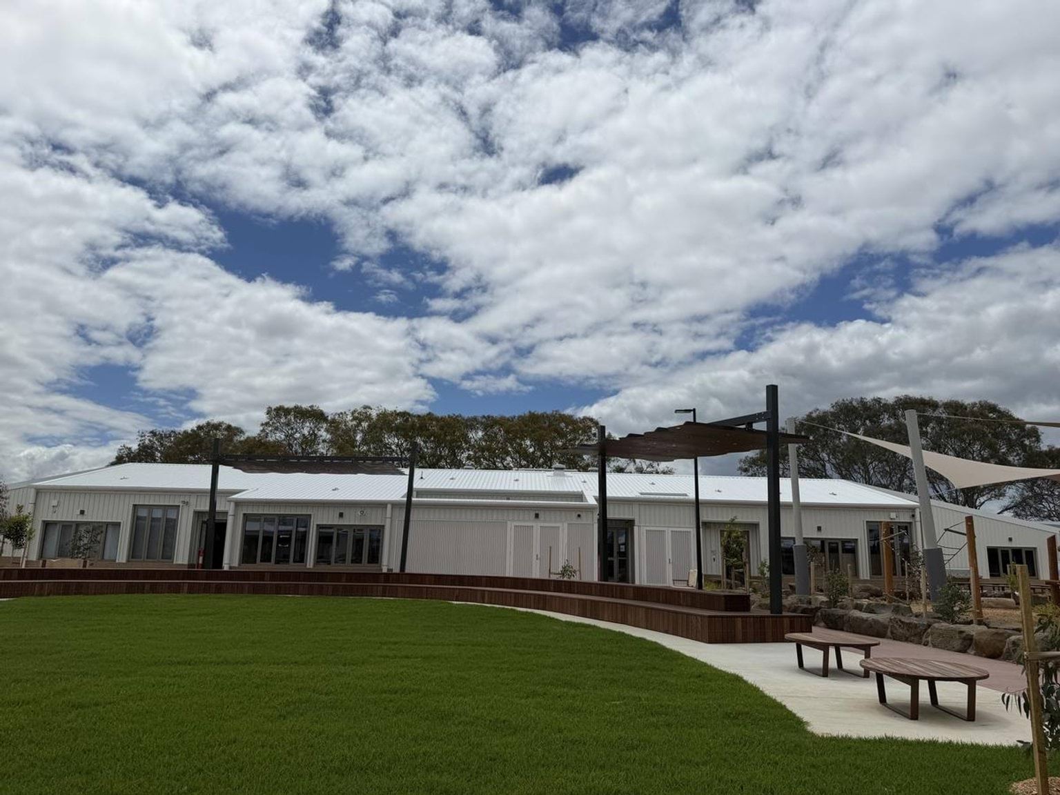 Outside Biyala Primary School. There is green grass in the foreground, wooden bench seating curving around it and a white building in the background. 
