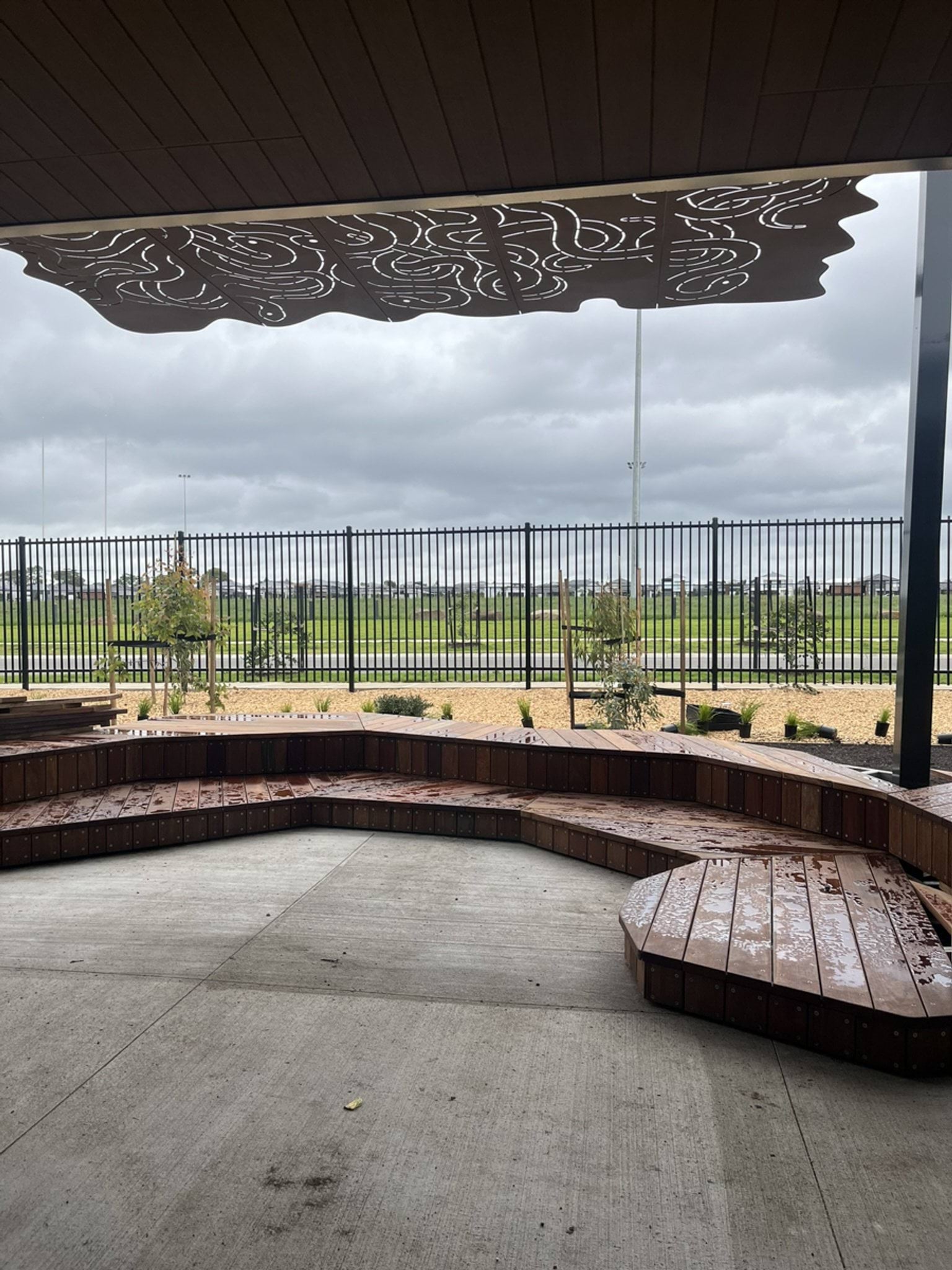A covered seating area outside, with a view of a fence, then a road and a green grassy area across the road.