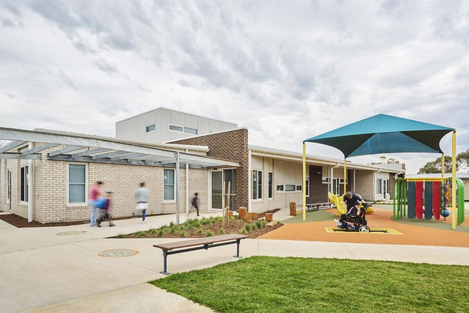 The exterior of a modern school building with cream and brown brick walls. A covered walkway runs along the front, and a play area with bright equipment and a shade sail is in the foreground.