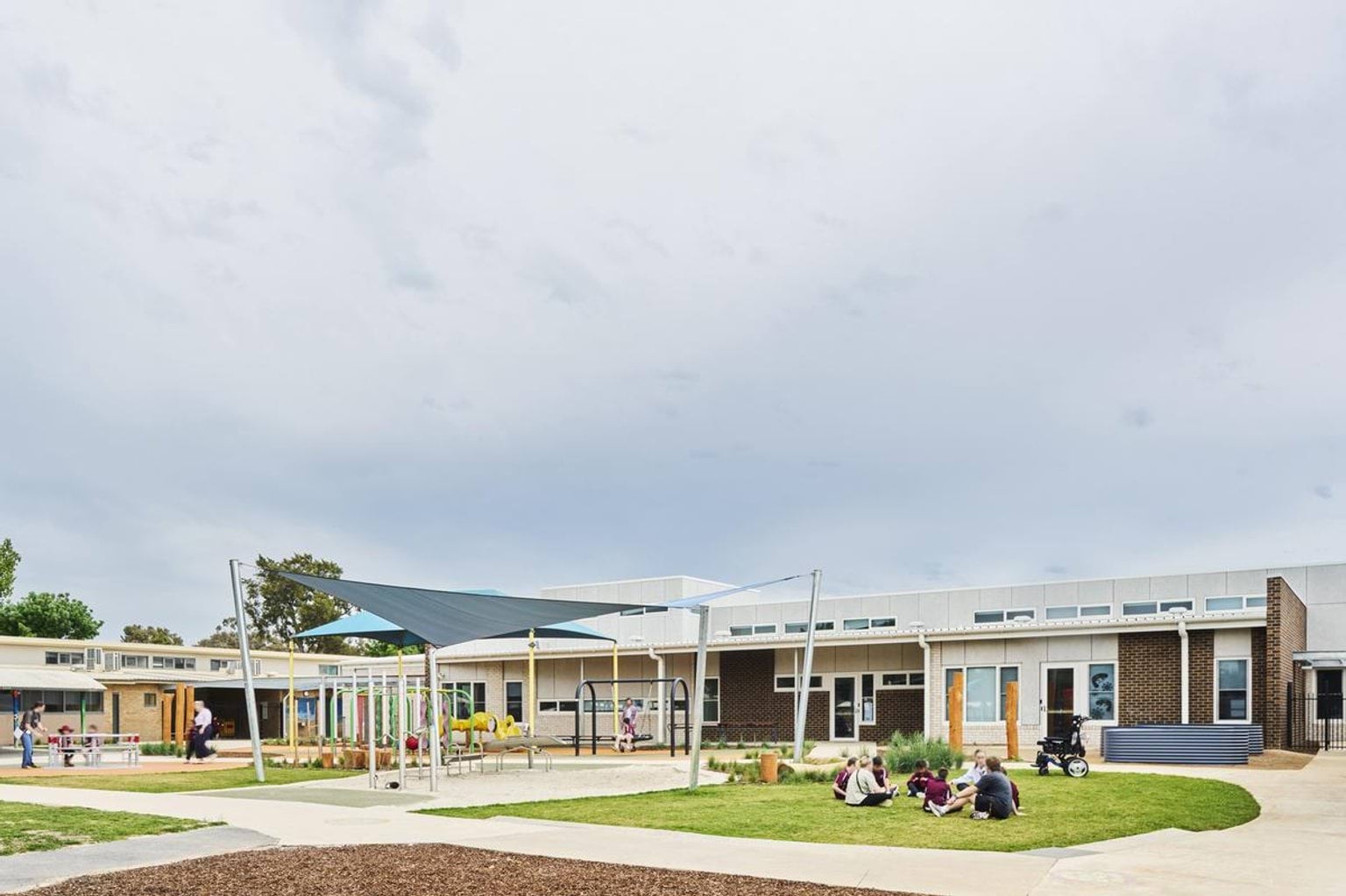A landscaped courtyard with grass, concrete paths, and a shade structure over a play area. The school building has cream and brown brick walls and large windows.