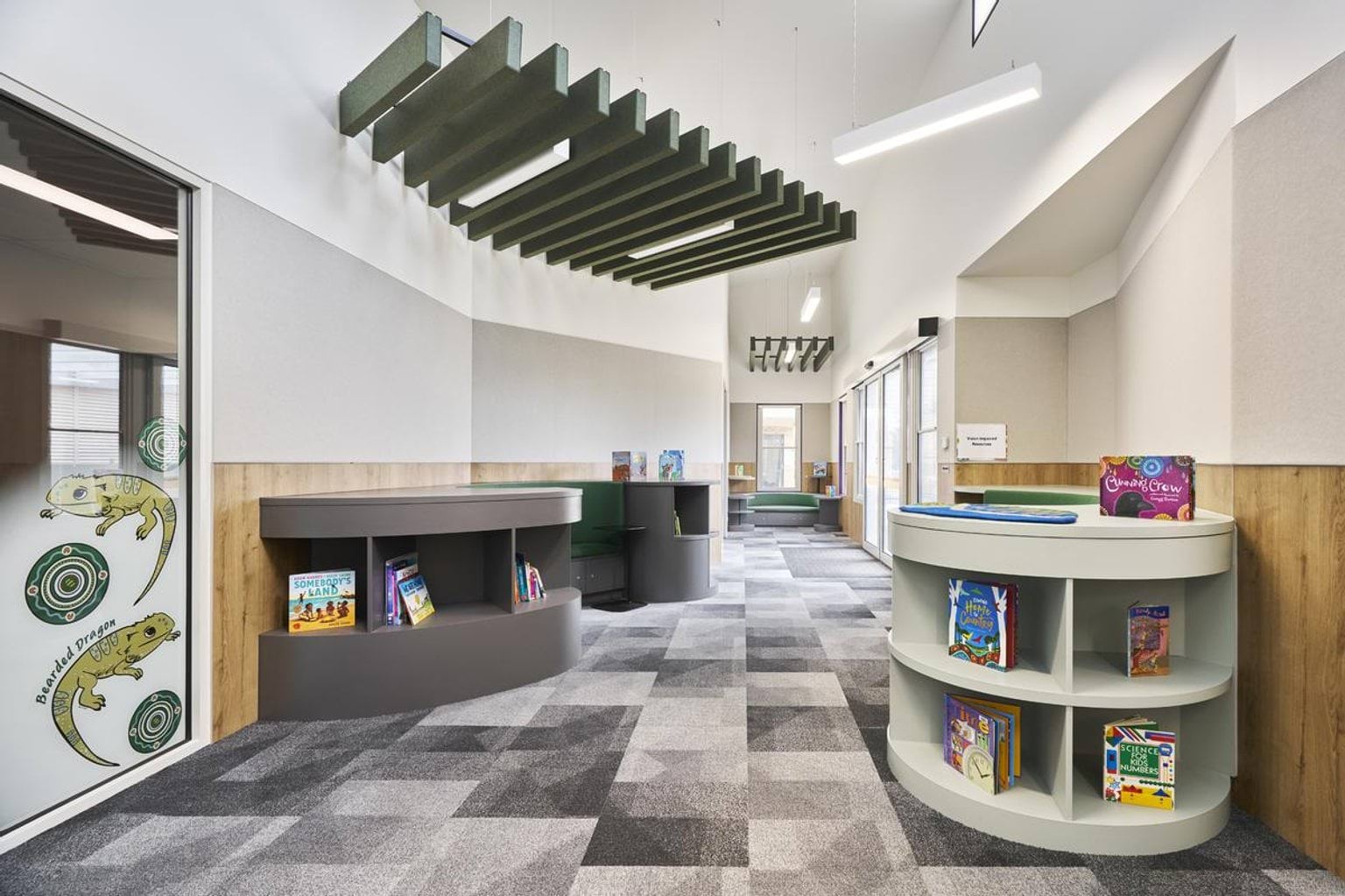 An open reading space with grey and green carpet, round shelving units filled with books, and timber wall panels. Green acoustic panels hang from the ceiling.