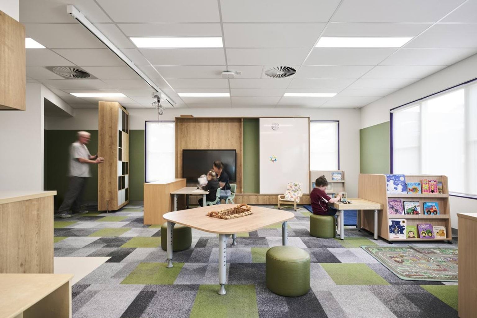 A classroom with patterned carpet, timber shelving, and activity tables. Bookshelves and a rug with colourful designs are arranged along the walls.
