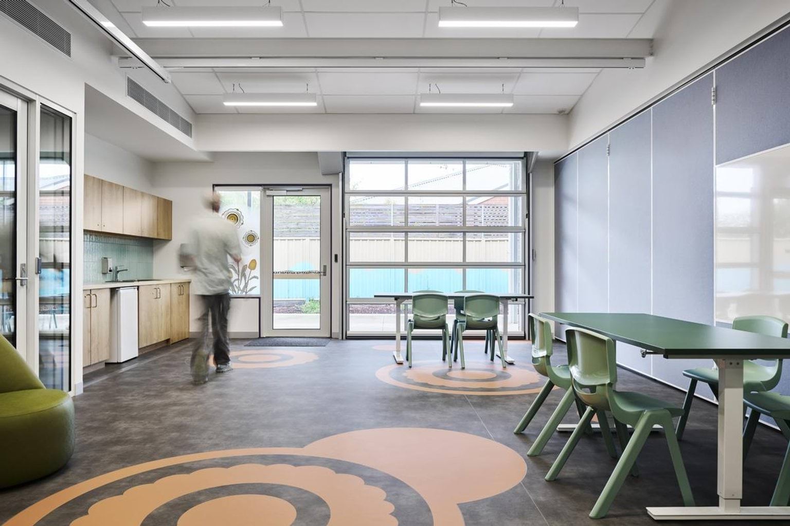 A bright classroom with a kitchenette, green tables, and patterned flooring featuring circular designs. Large windows and sliding doors open to an outdoor area.