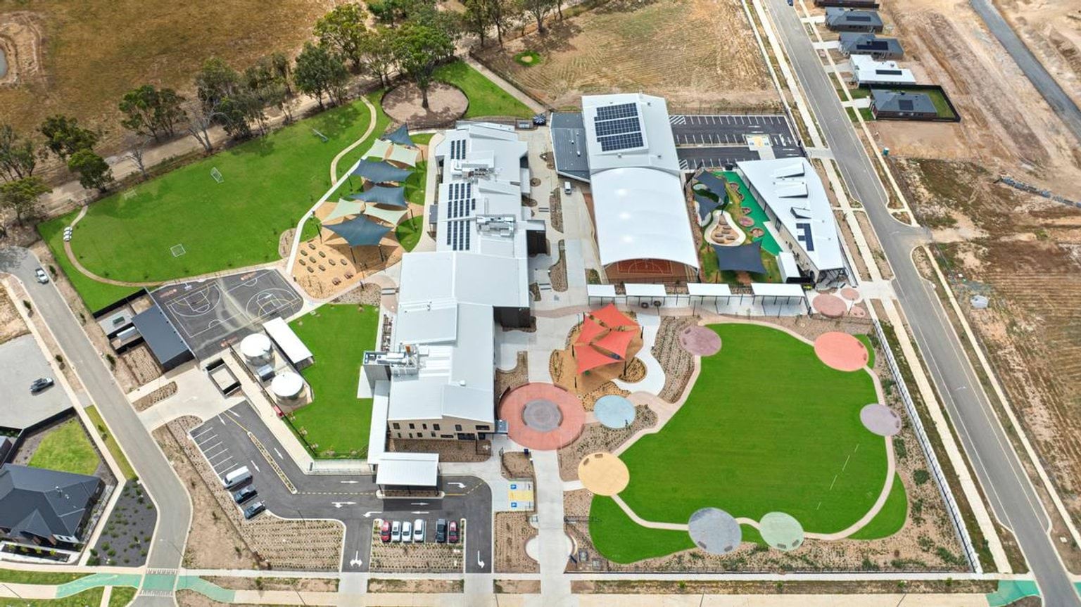 Aerial view of the school site showing new buildings, landscaped play areas, shade structures, and sports courts surrounded by green spaces
