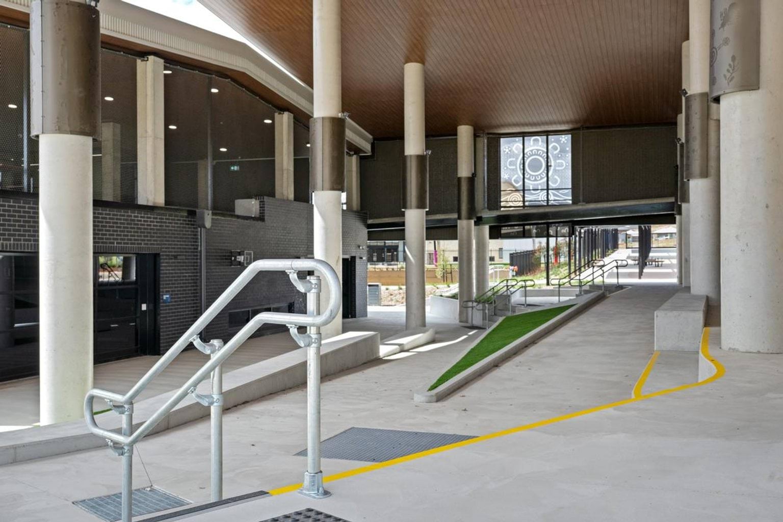Covered walkway under a school building with concrete ramps, stairs, and railings, designed for accessible movement between levels