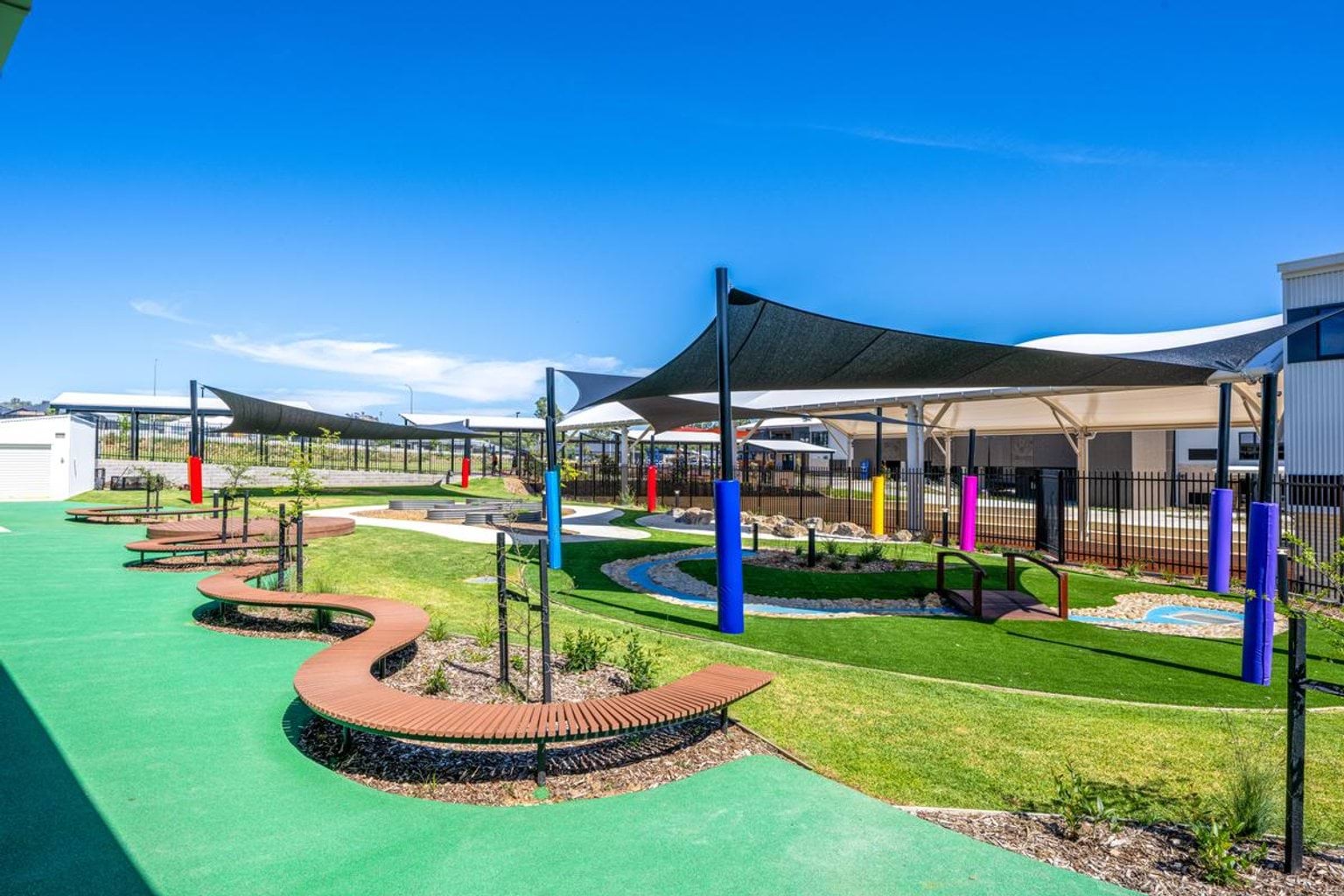 Outdoor learning space with shade sails, colourful posts, curved seating, and green play surfaces near school buildings