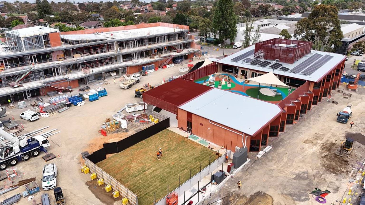 Aerial view of the kindergarten building with solar panels on the roof and a landscaped play area featuring shade sails and bright surfaces.