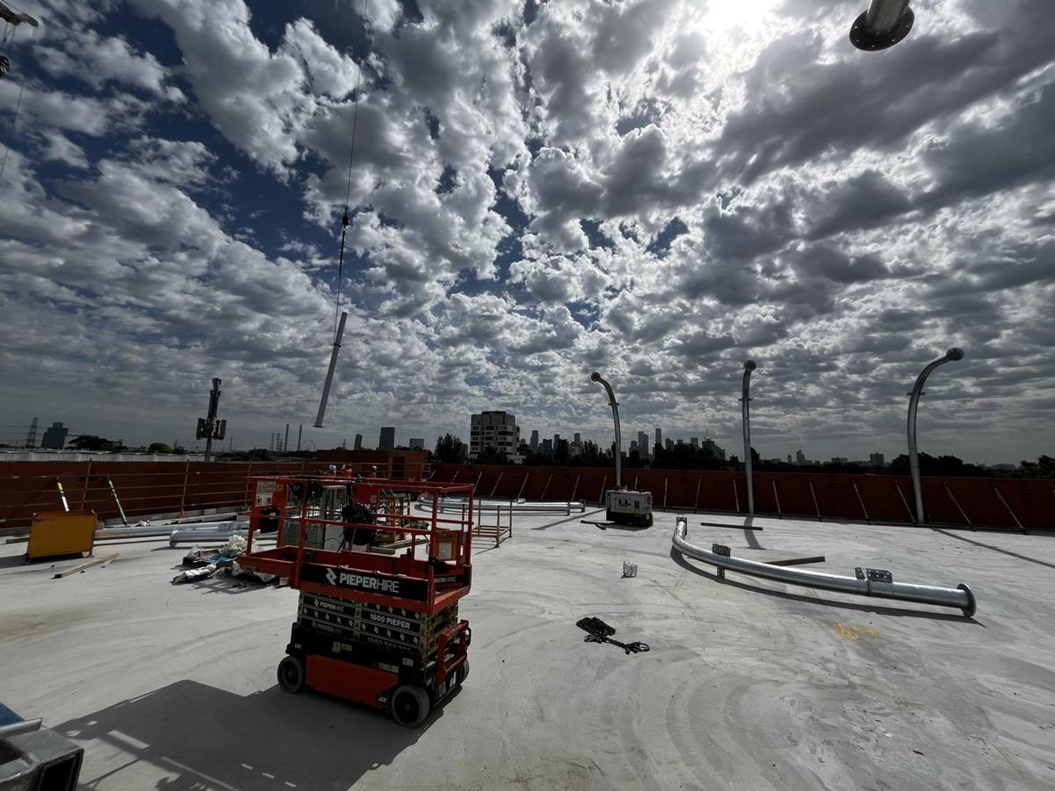 A rooftop construction area with curved metal frames and scattered building materials under a cloudy sky. A scissor lift is in the foreground.
