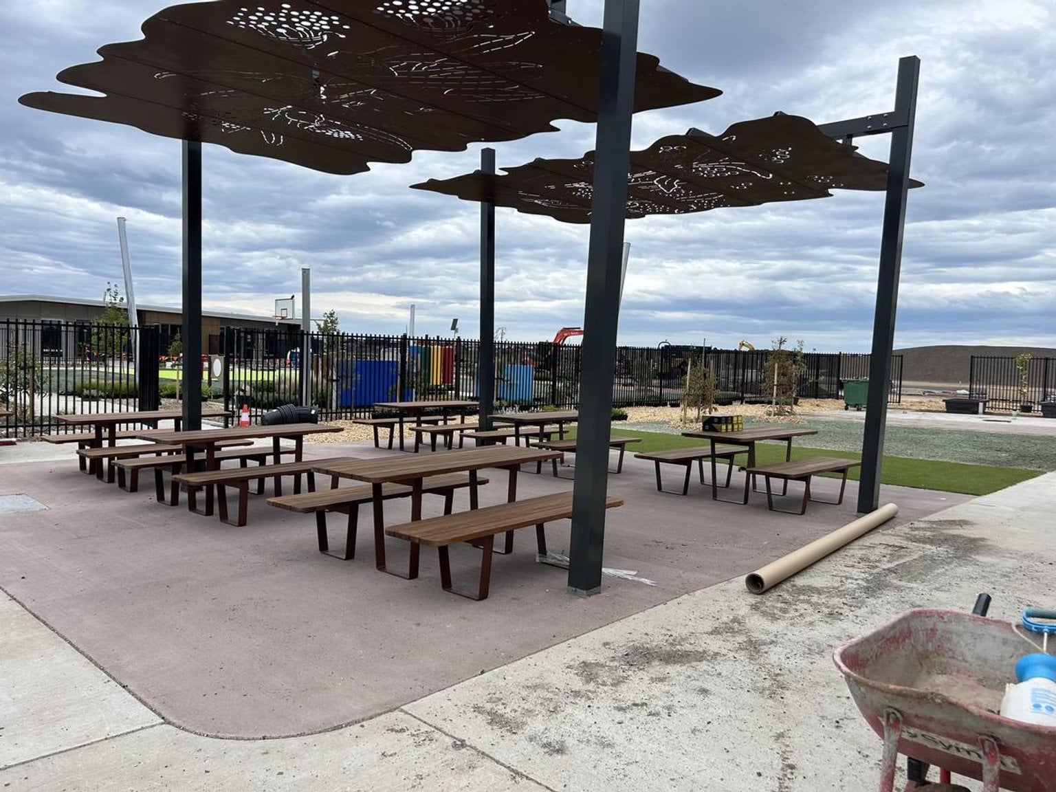 A courtyard with picnic tables on artificial grass under a decorative metal shade structure, surrounded by landscaped garden beds.