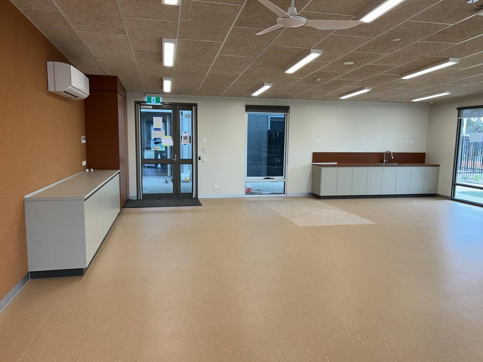 A large classroom with light flooring, built-in cabinetry, and multiple doors leading outside, under bright ceiling lights.