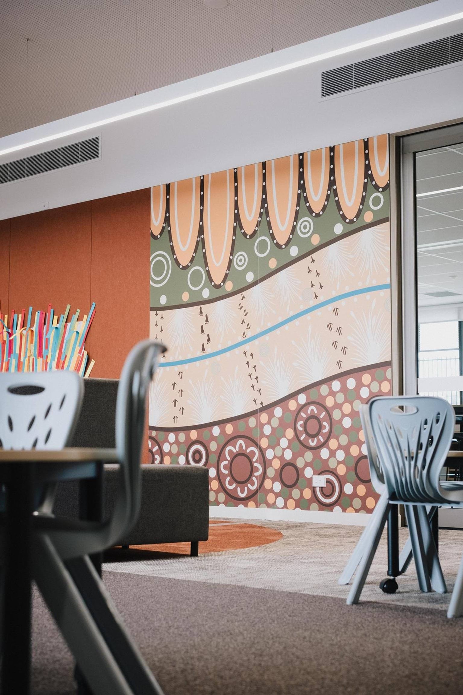 Interior learning space with a large Aboriginal artwork mural featuring earthy tones and circular patterns, with chairs and tables in the foreground