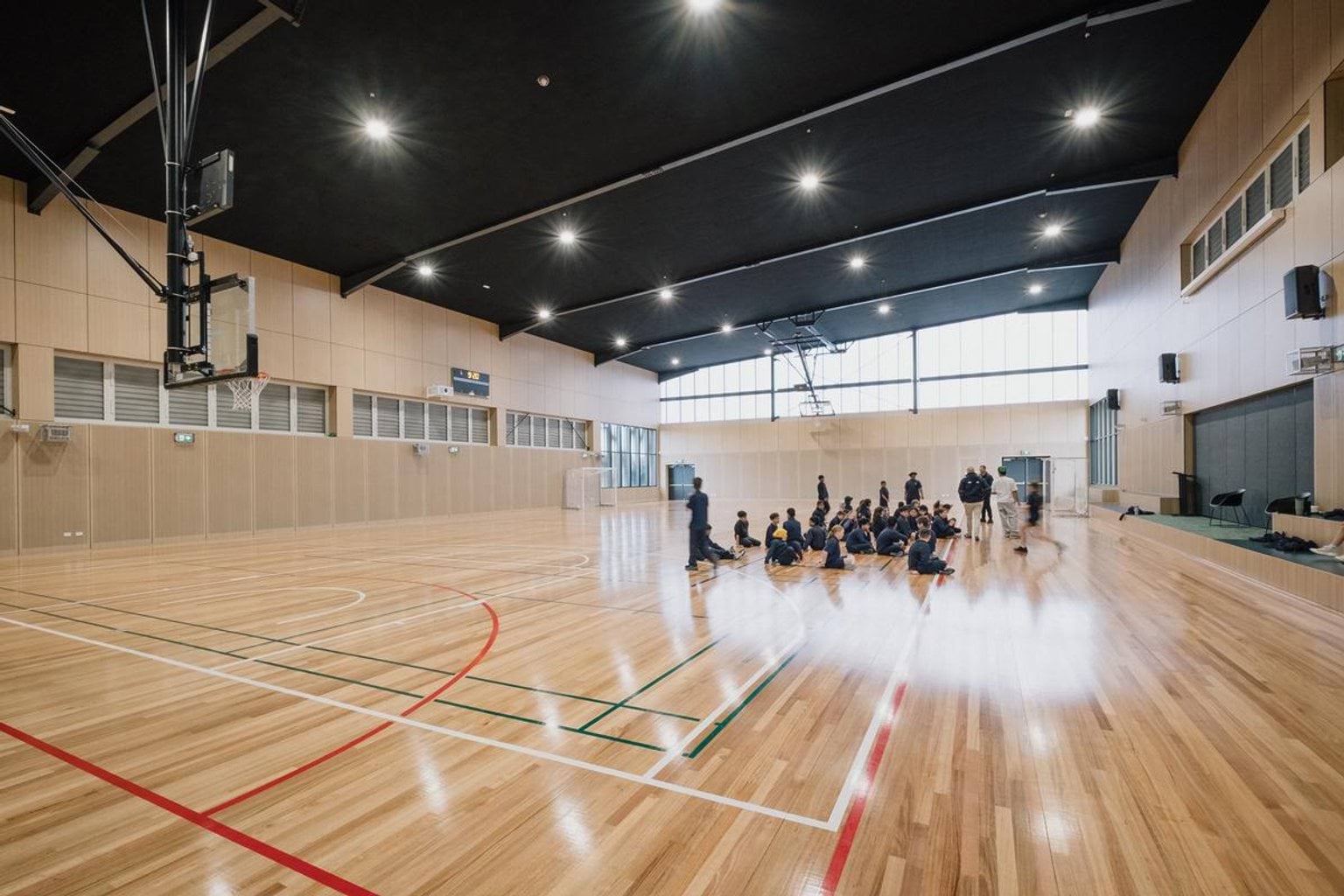 Indoor gymnasium with timber flooring, basketball hoops and students seated in a group on the court
