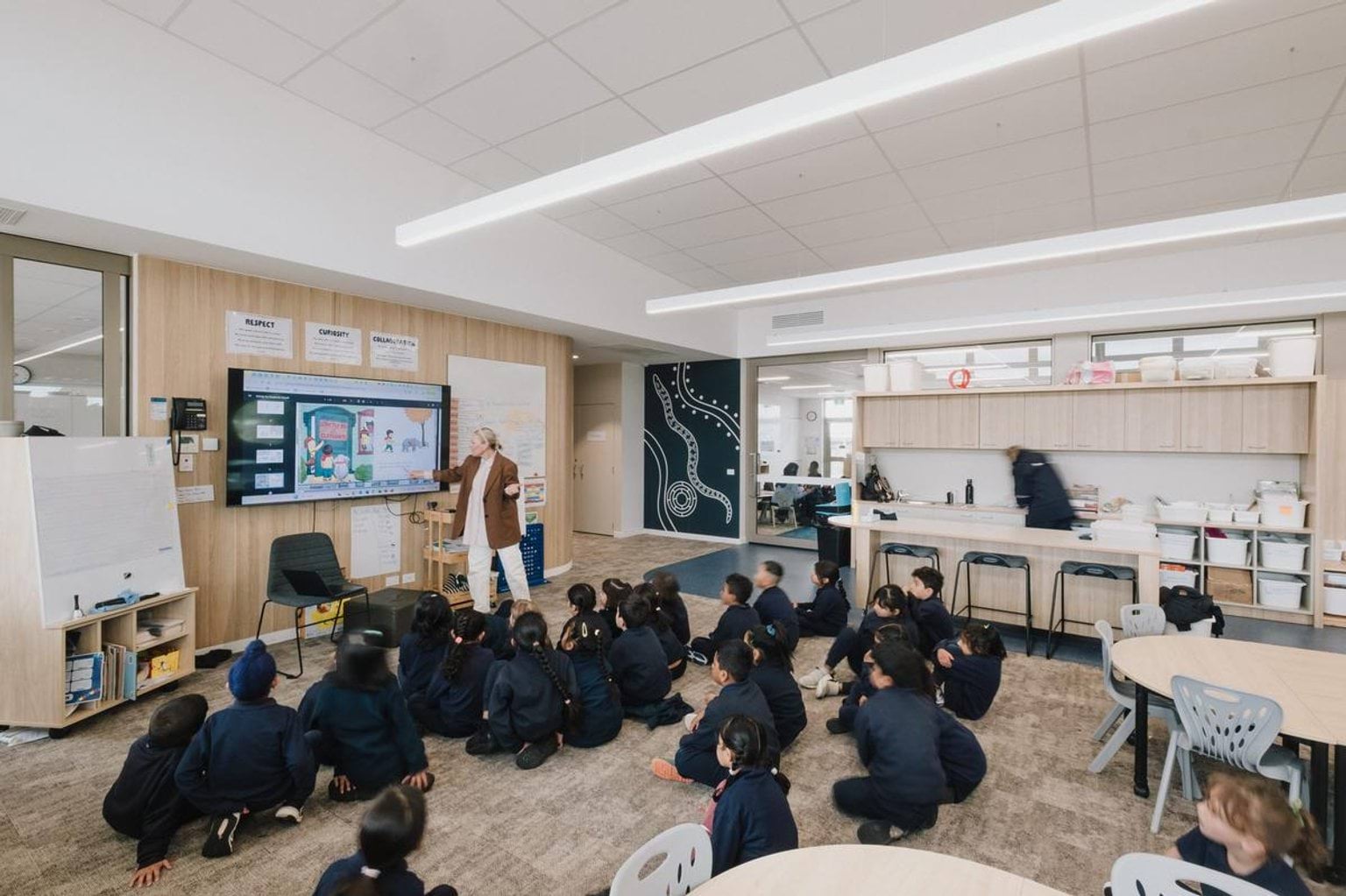 Classroom with students seated on carpet facing interactive screen and teacher presenting at the front
