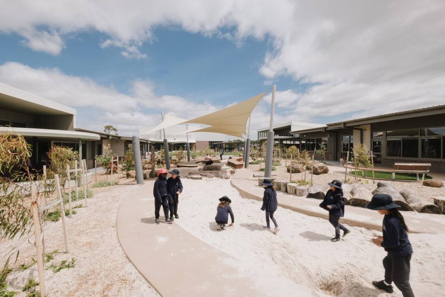 Outdoor play area with shade sails, sandpit and natural landscaping, with children playing along the path