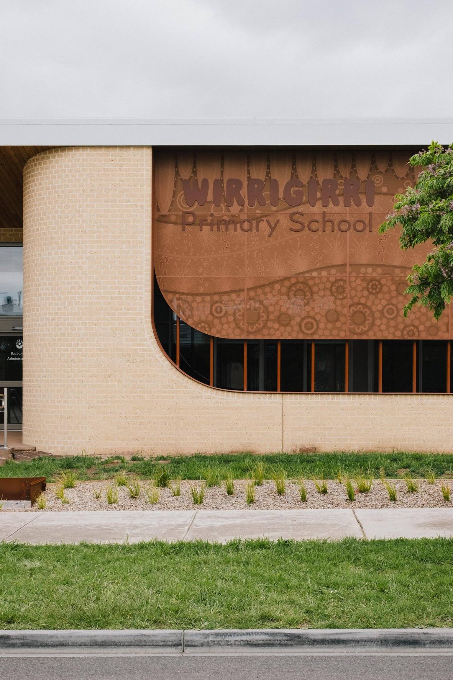 Exterior view of Wirrigirri Primary School building with curved brick walls, patterned facade displaying the school name, and landscaped garden in front