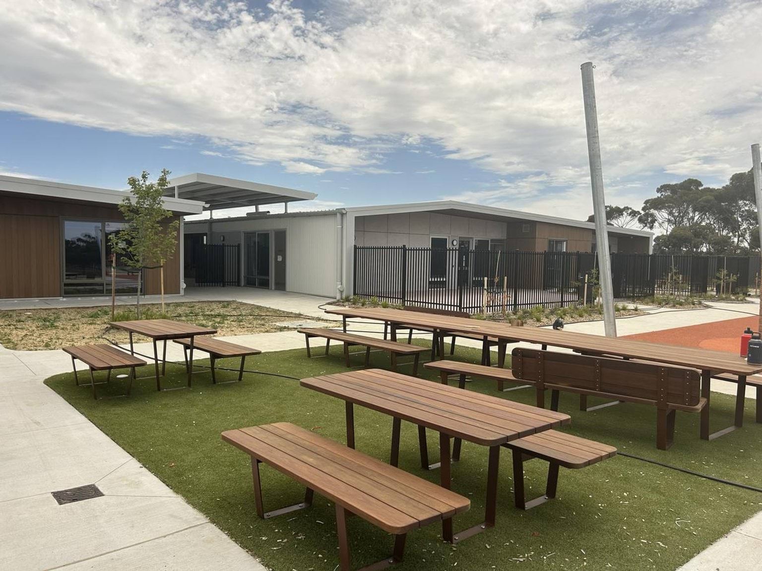 Wooden picnic tables arranged on artificial grass in a courtyard between modern school buildings with timber and grey cladding.