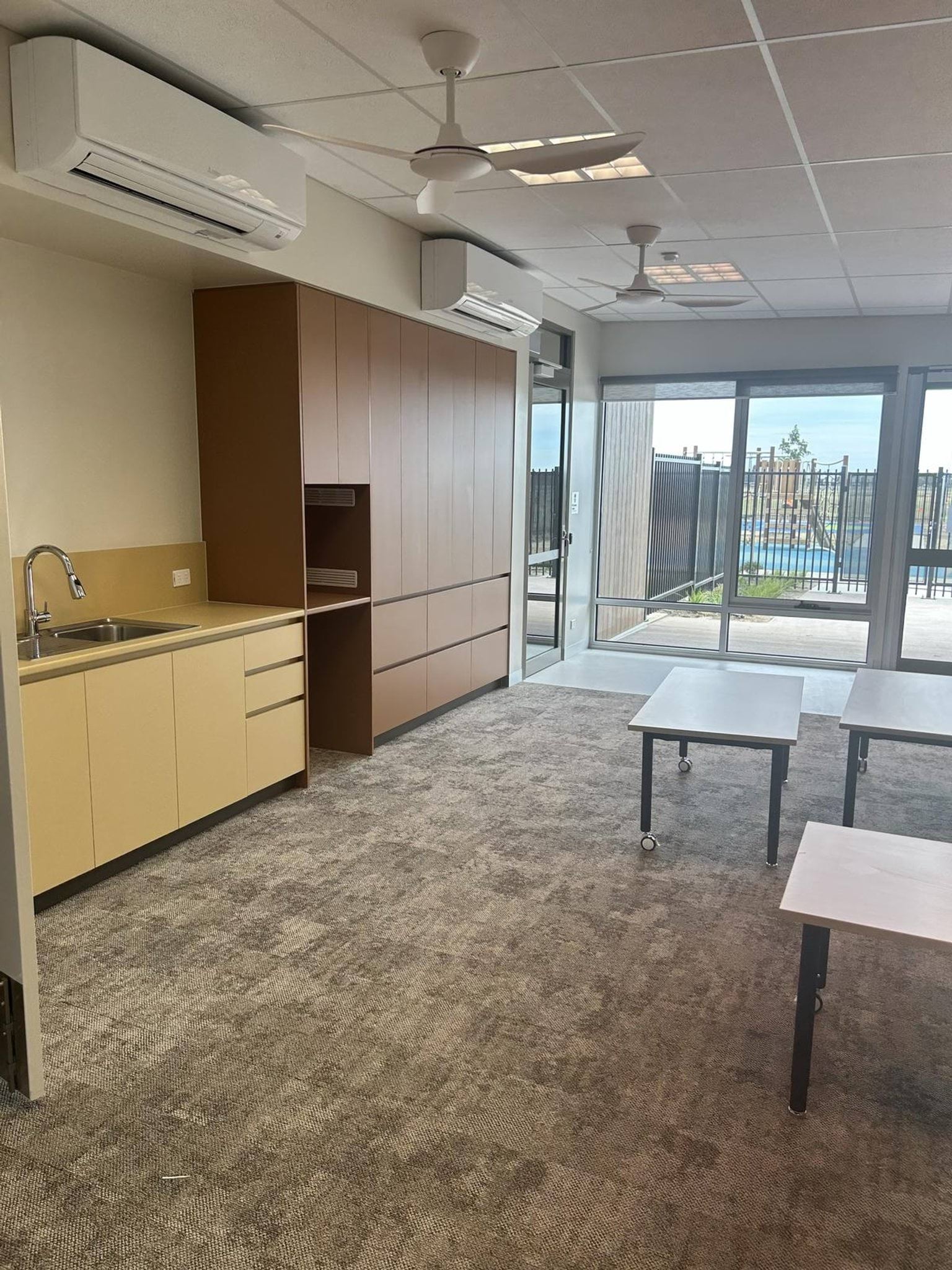A classroom with carpet flooring, built-in cabinetry, a small sink, and large windows opening to an outdoor area.
