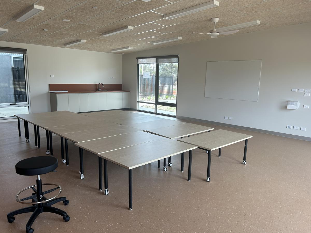 A classroom with modular tables arranged in a square, a stool, and a small sink area near large windows.
