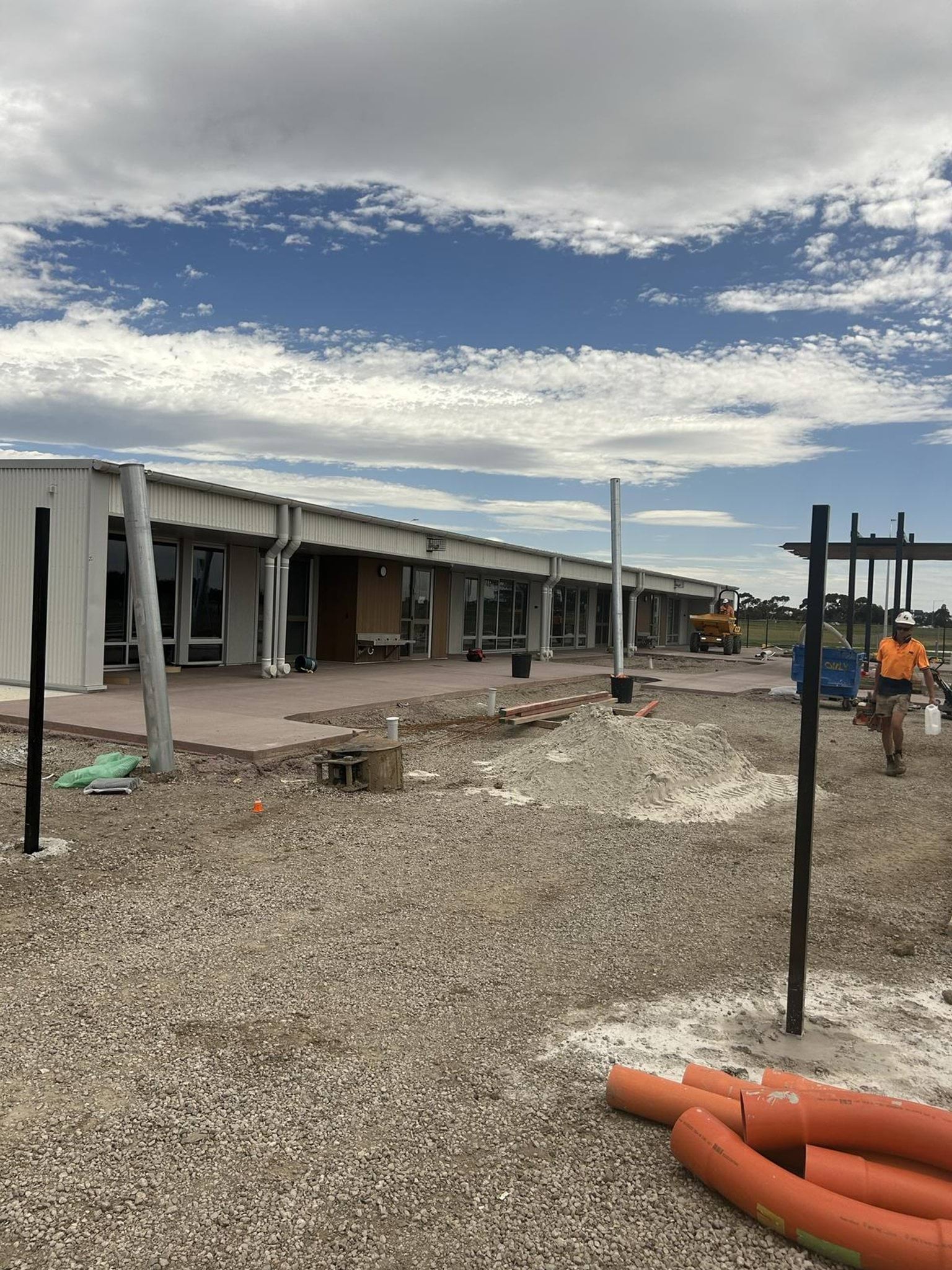 A single-storey kindergarten building with a covered walkway and glass doors. The foreground has gravel, orange pipes, and metal posts as part of ongoing landscaping work.