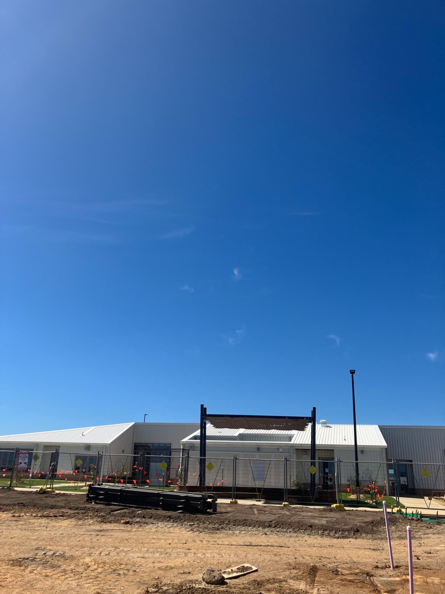 The front of a school building with white roofing and temporary fencing. Bare soil and construction materials are visible in the foreground.