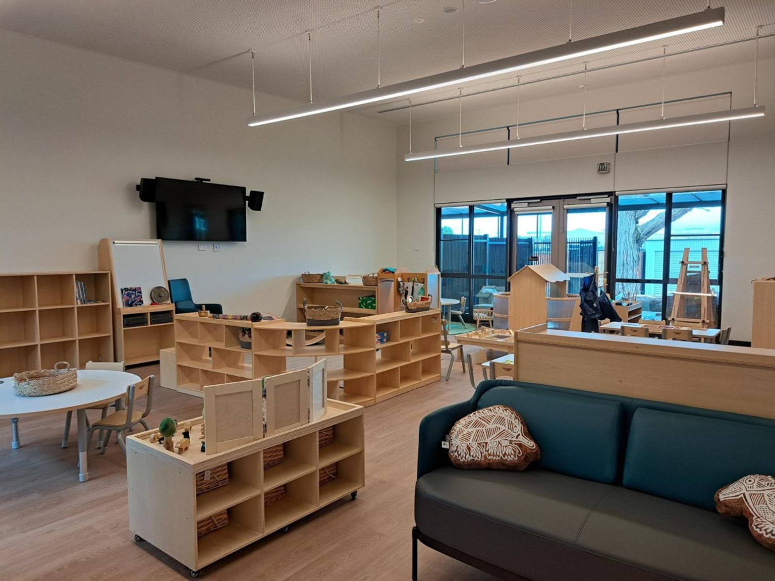 A kindergarten learning room with timber shelves, small tables and chairs, a TV on the wall, and large windows facing the outdoor play area.