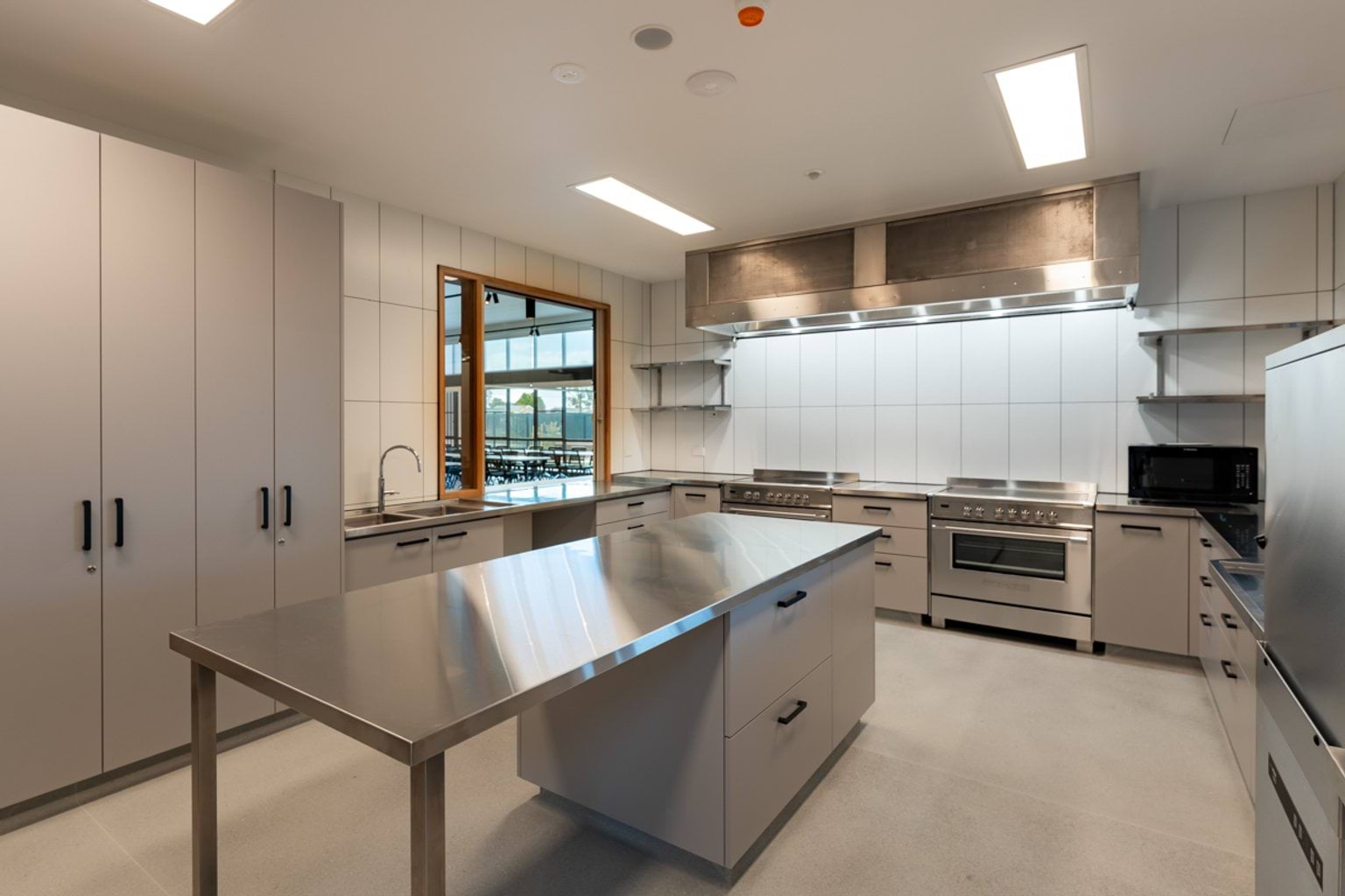 A commercial-style kitchen with stainless steel benches, multiple ovens, and white tiled walls.