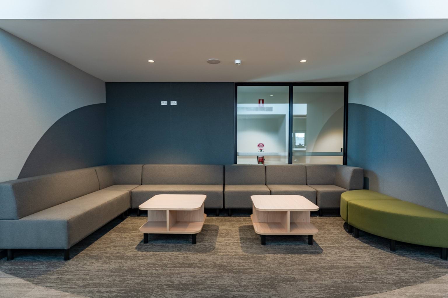 A lounge-style space with grey sofas, a green bench seat, and timber tables against a feature wall.