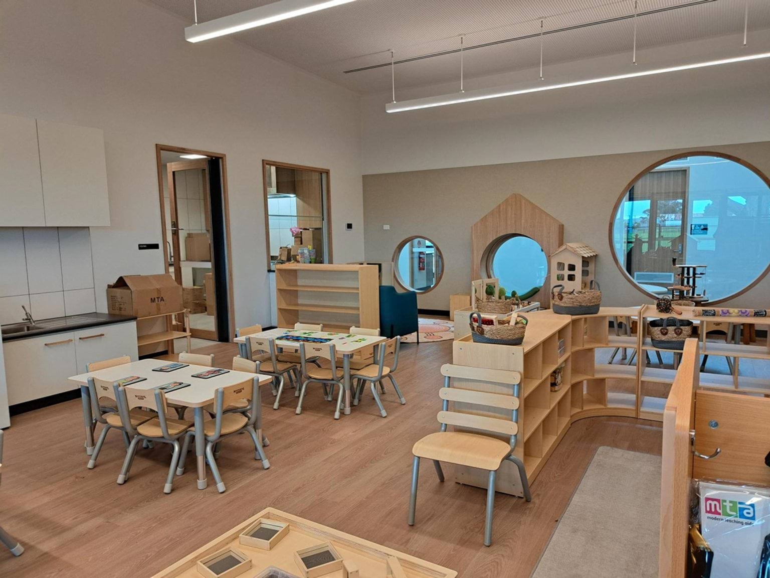 A kindergarten classroom with small tables, timber shelving, circular feature windows, and play equipment arranged around the room.