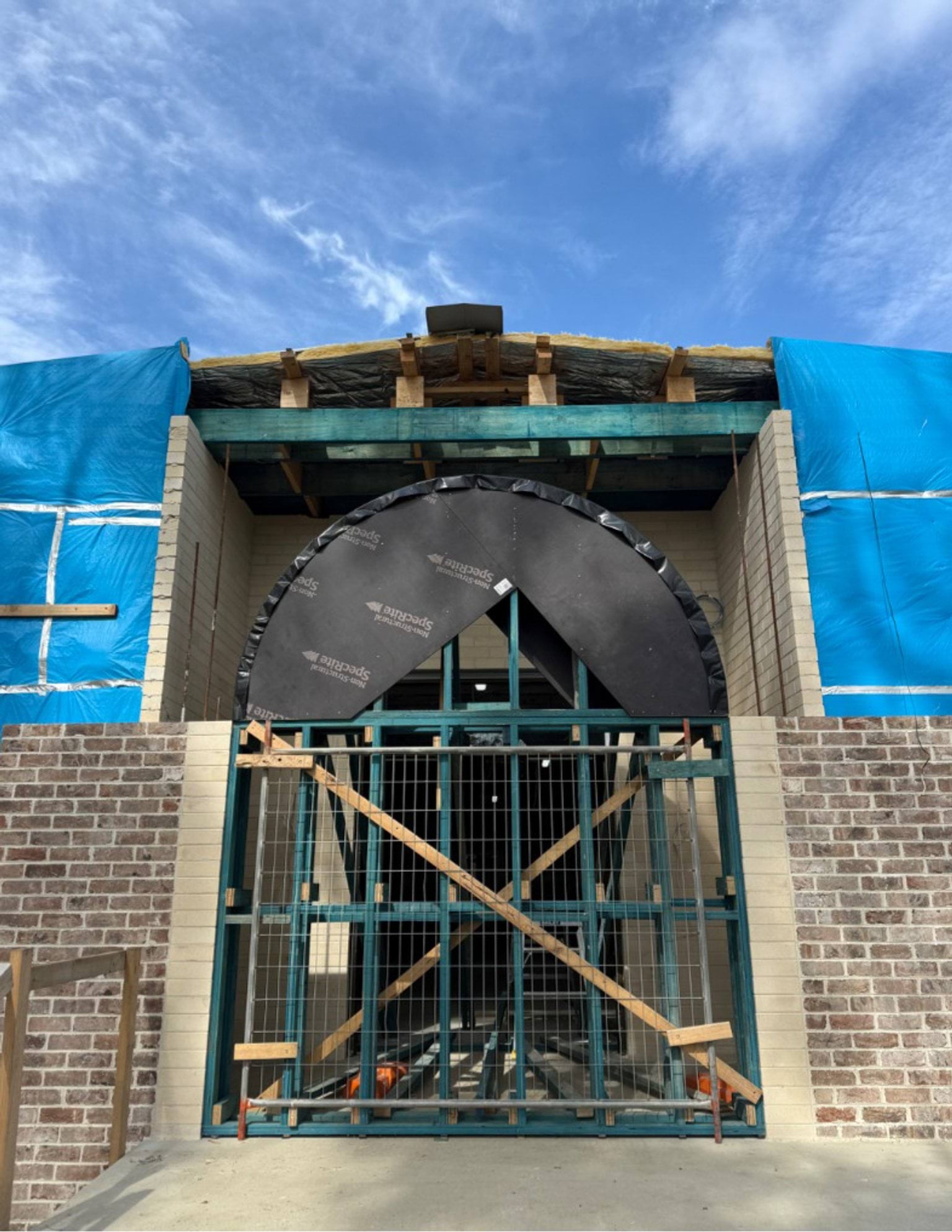 A partially built school entryway with a circular arch frame, blue protective sheeting, and exposed roof timbers.