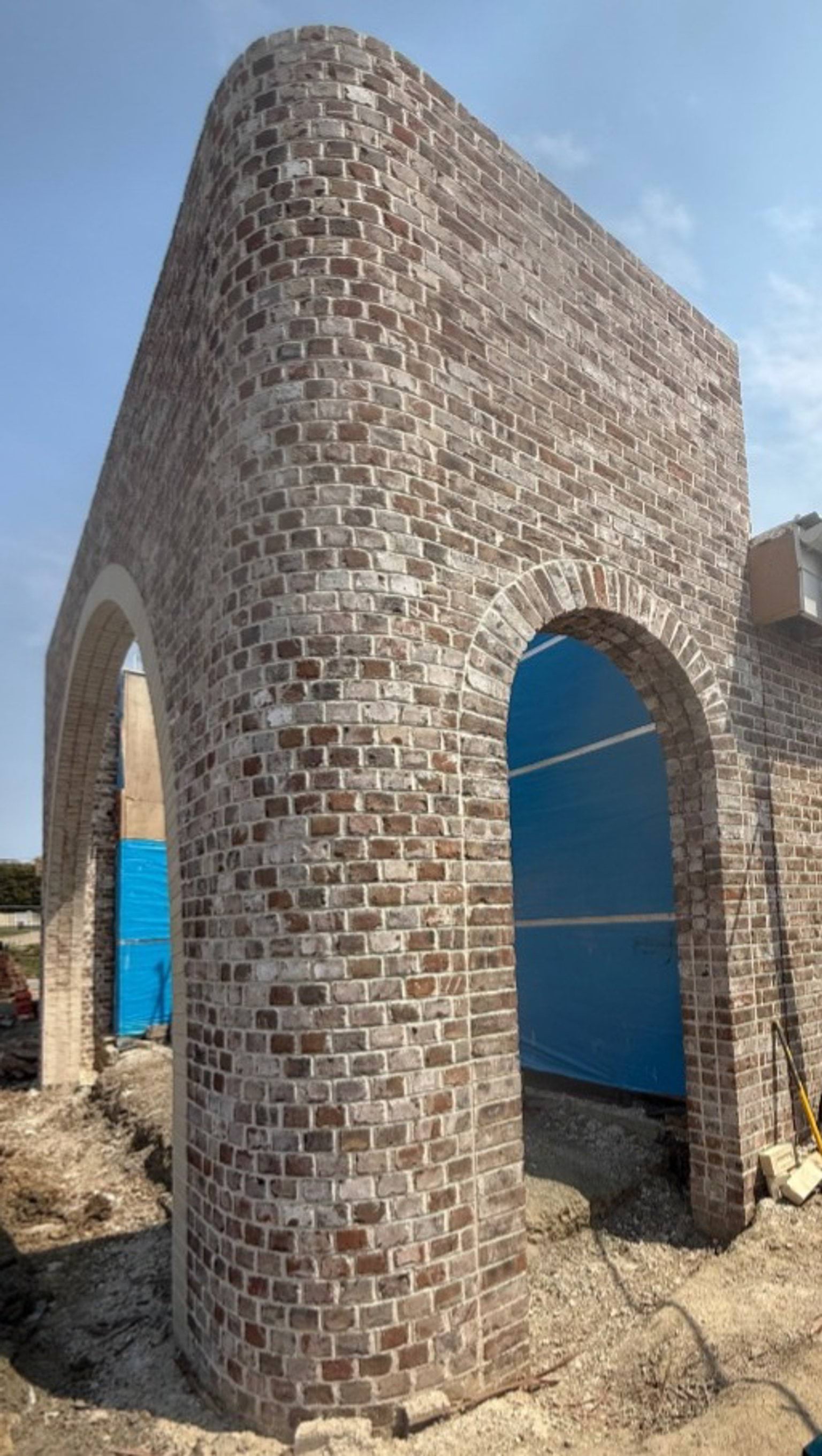 A curved brick archway under construction, with exposed ground and temporary blue sheeting behind the structure.