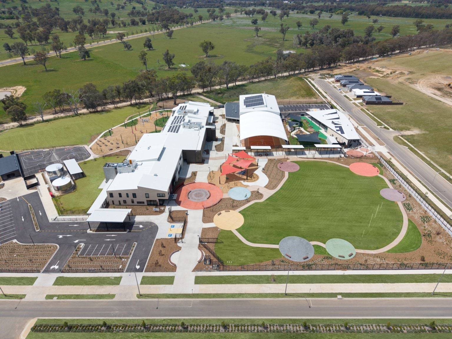 Aerial view of the completed school campus with buildings, sports courts, landscaped play areas and surrounding open land.