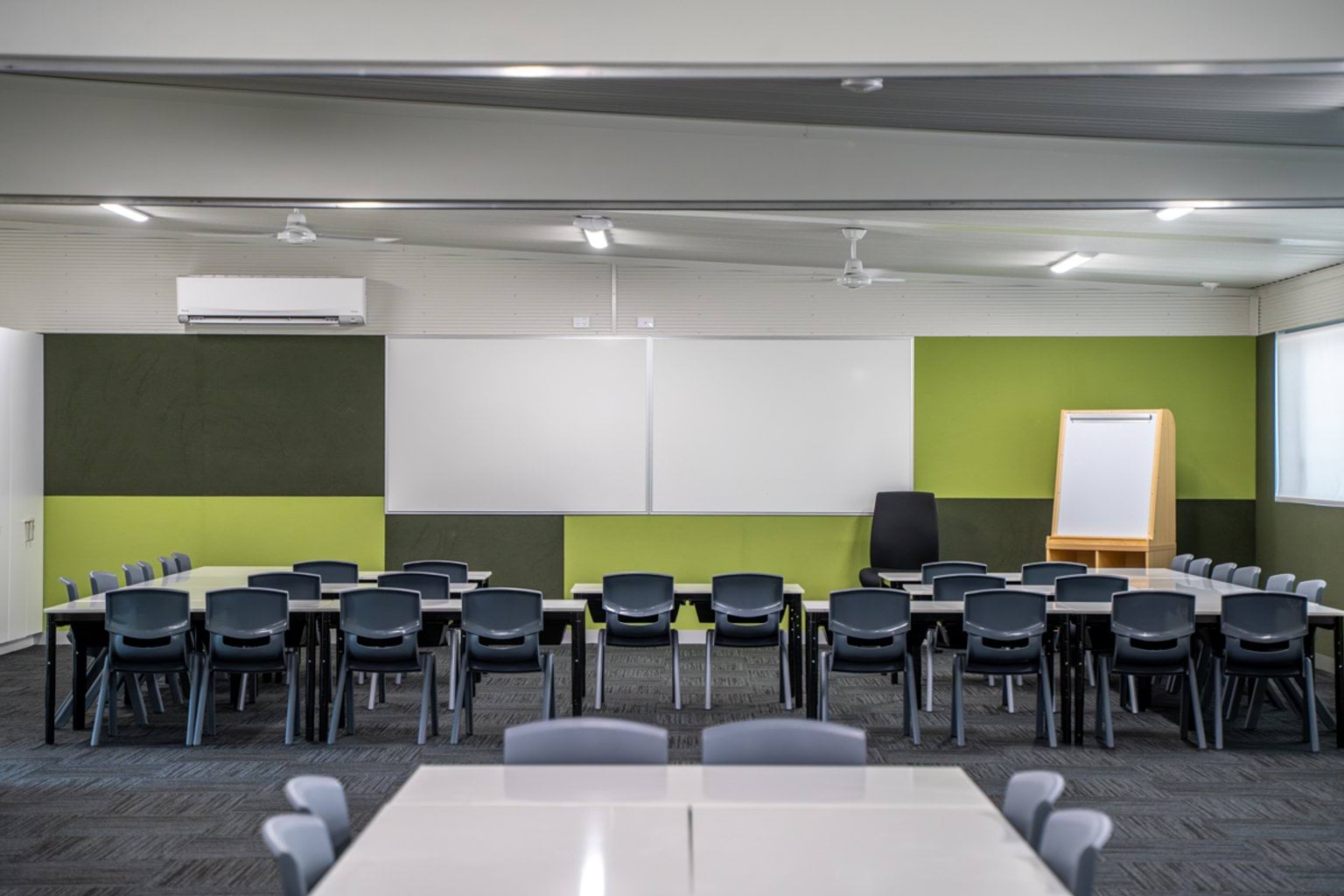 A modular classroom with green and white walls, ceiling fans, and rows of desks facing large whiteboards.