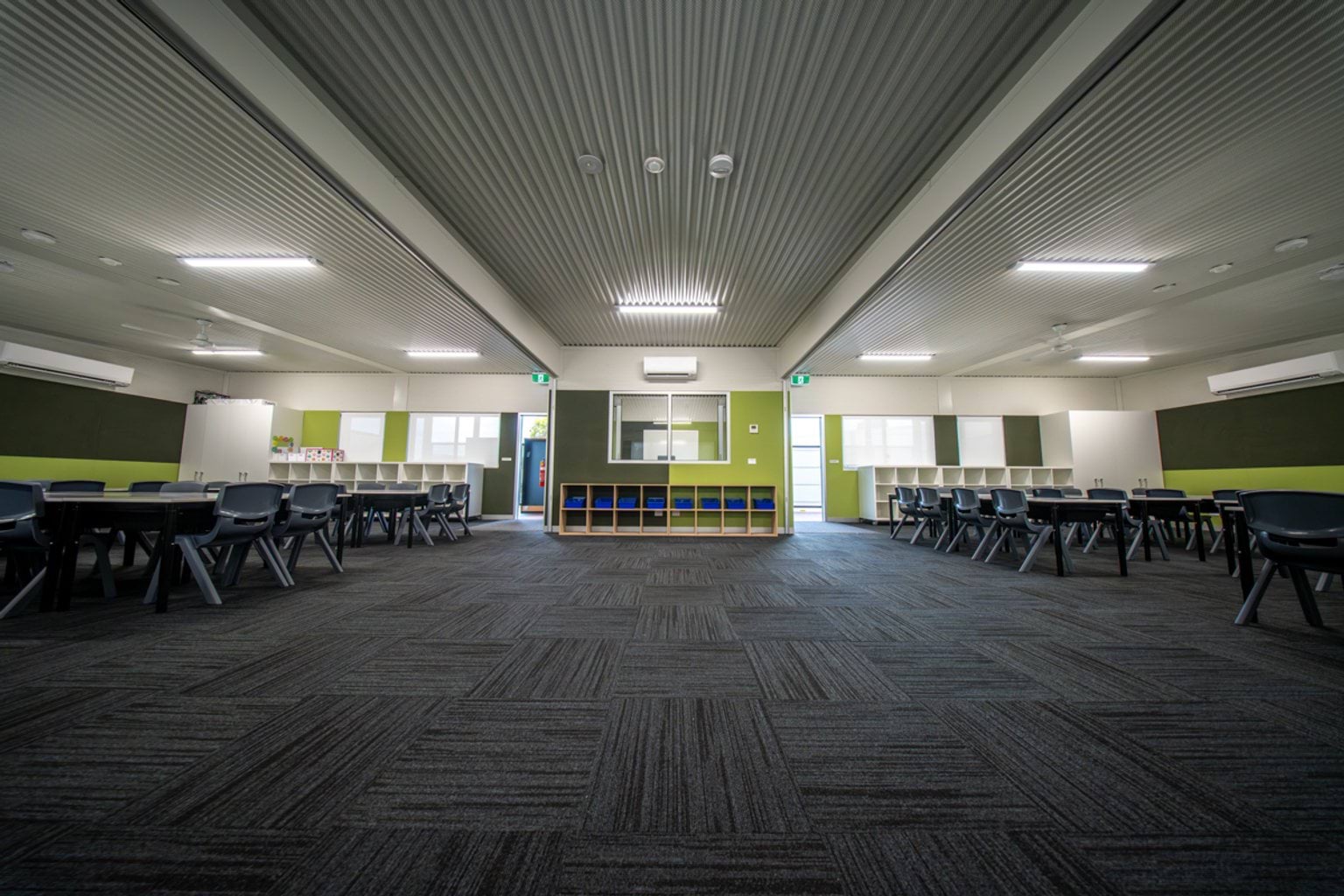A spacious modular classroom with carpet tiles, green feature walls, and rows of tables and chairs arranged along both sides.