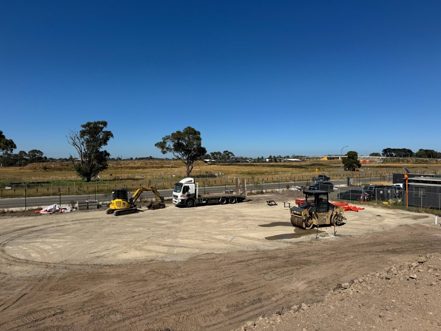 Construction vehicles working on a cleared site with compacted soil, beside a fenced road and open paddocks.