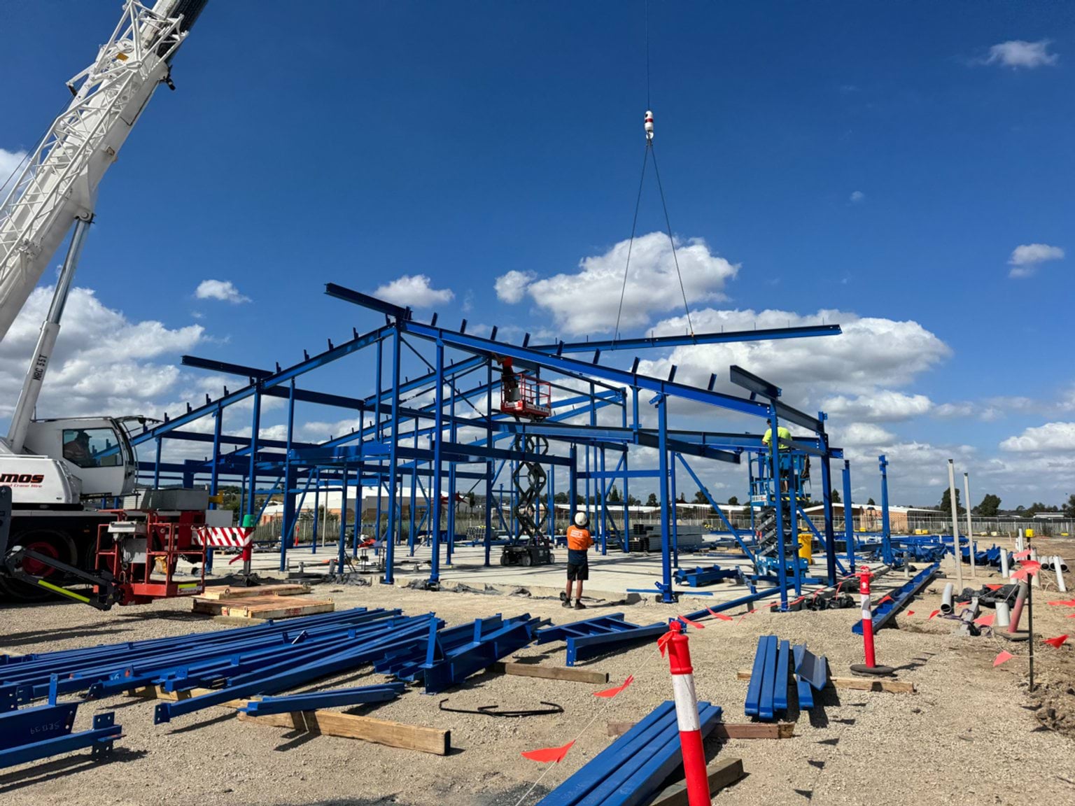 A steel frame being assembled on a concrete slab, with a crane and workers guiding sections into place.