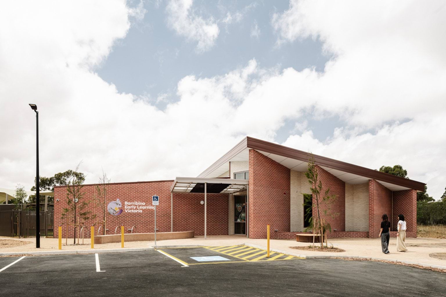 A school building with tall red brick columns and a landscaped area, with two people walking along the path.