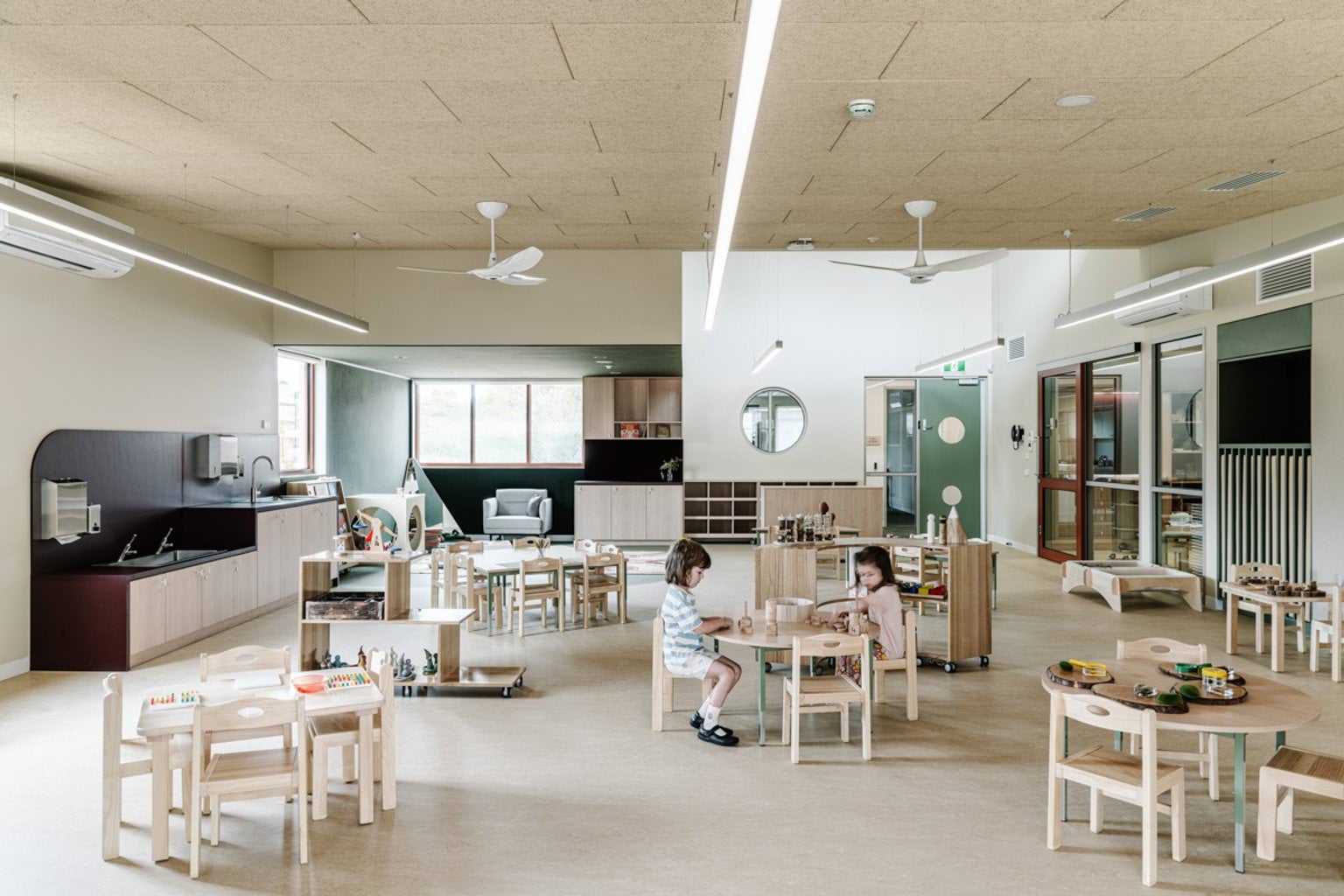 A large kindergarten classroom with small tables, timber play furniture, and activity stations spread across the room.