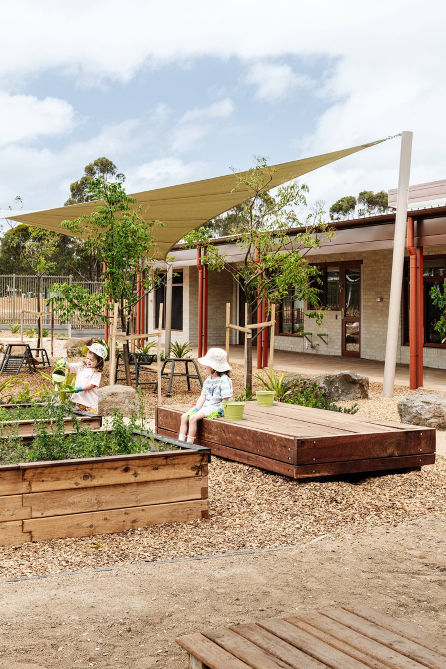 Children exploring raised garden beds and natural play elements in an outdoor learning area with shade sails.