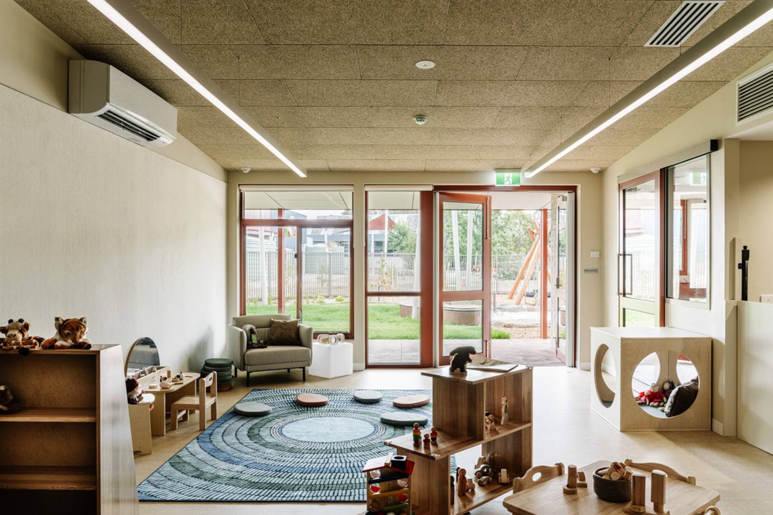 An indoor playroom with natural timber furniture, soft seating, and a round blue patterned rug, opening to an outdoor play space.