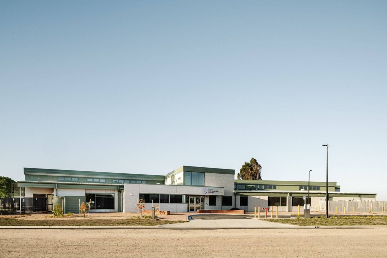 A wide exterior view of the school building with large windows, landscaped beds, and an open forecourt.