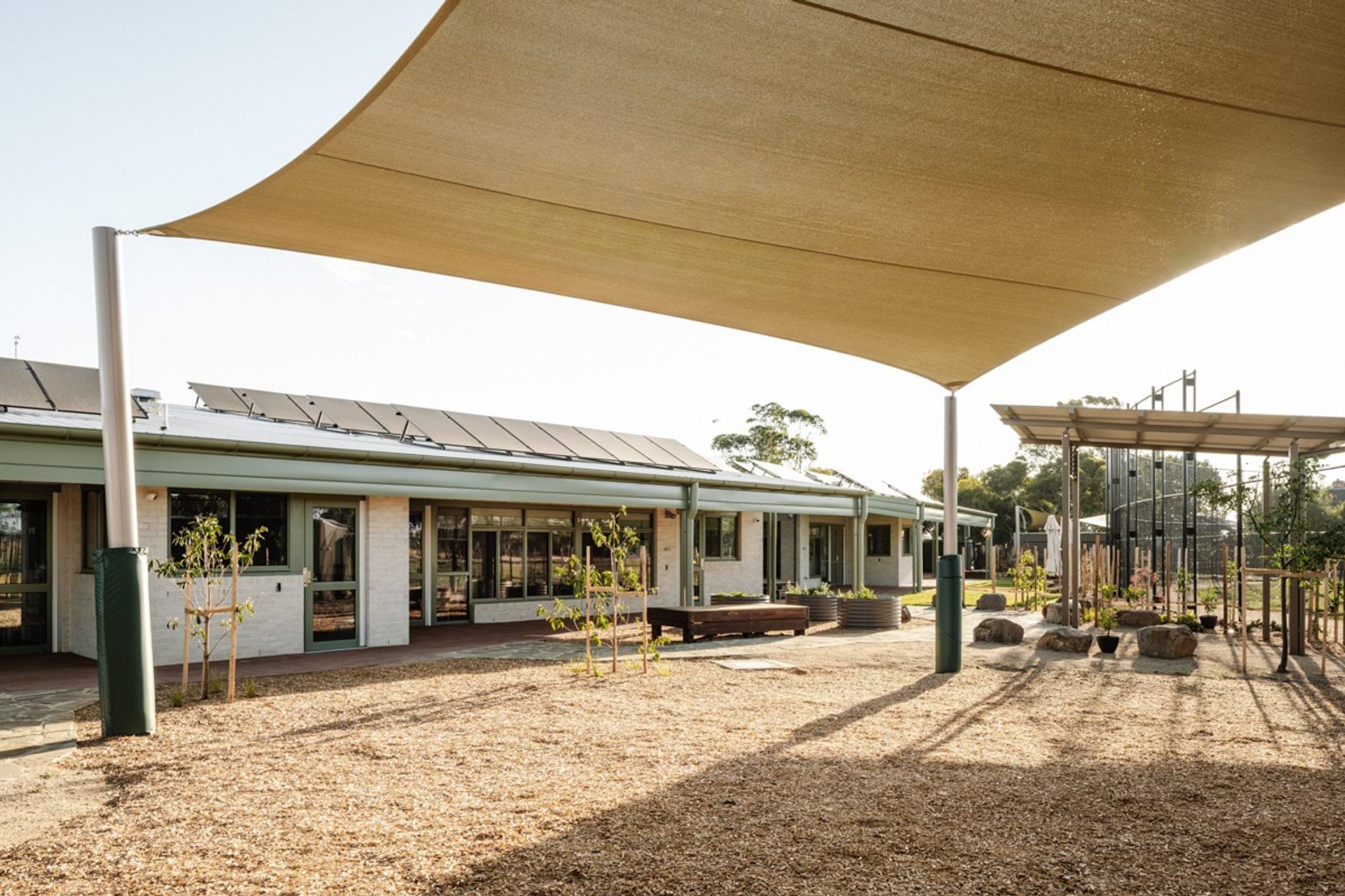The school building viewed from the outdoor play space, shaded by large sails and surrounded by young plantings.