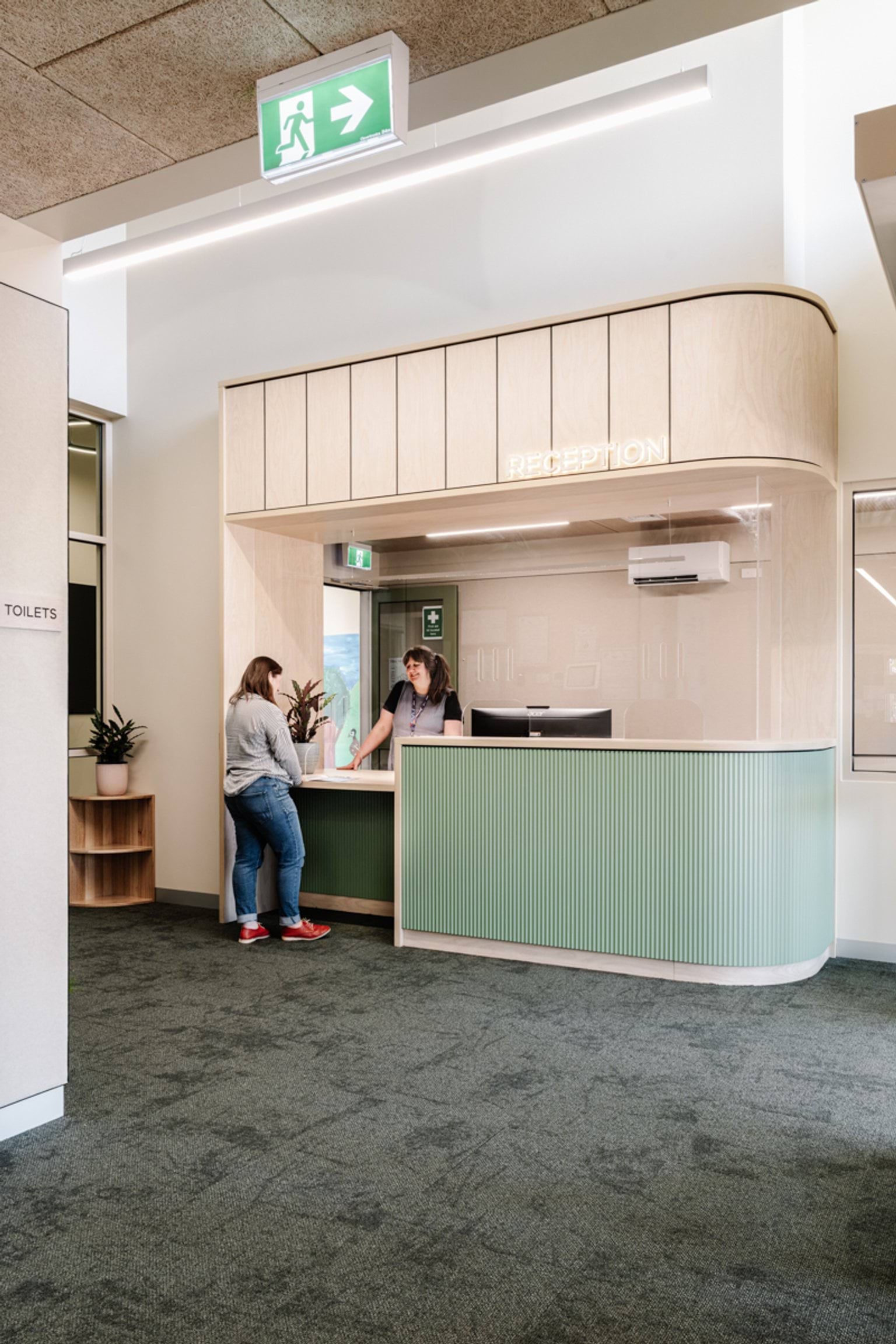 A school reception area with a curved timber and green counter, where a person is speaking with staff behind the desk.
