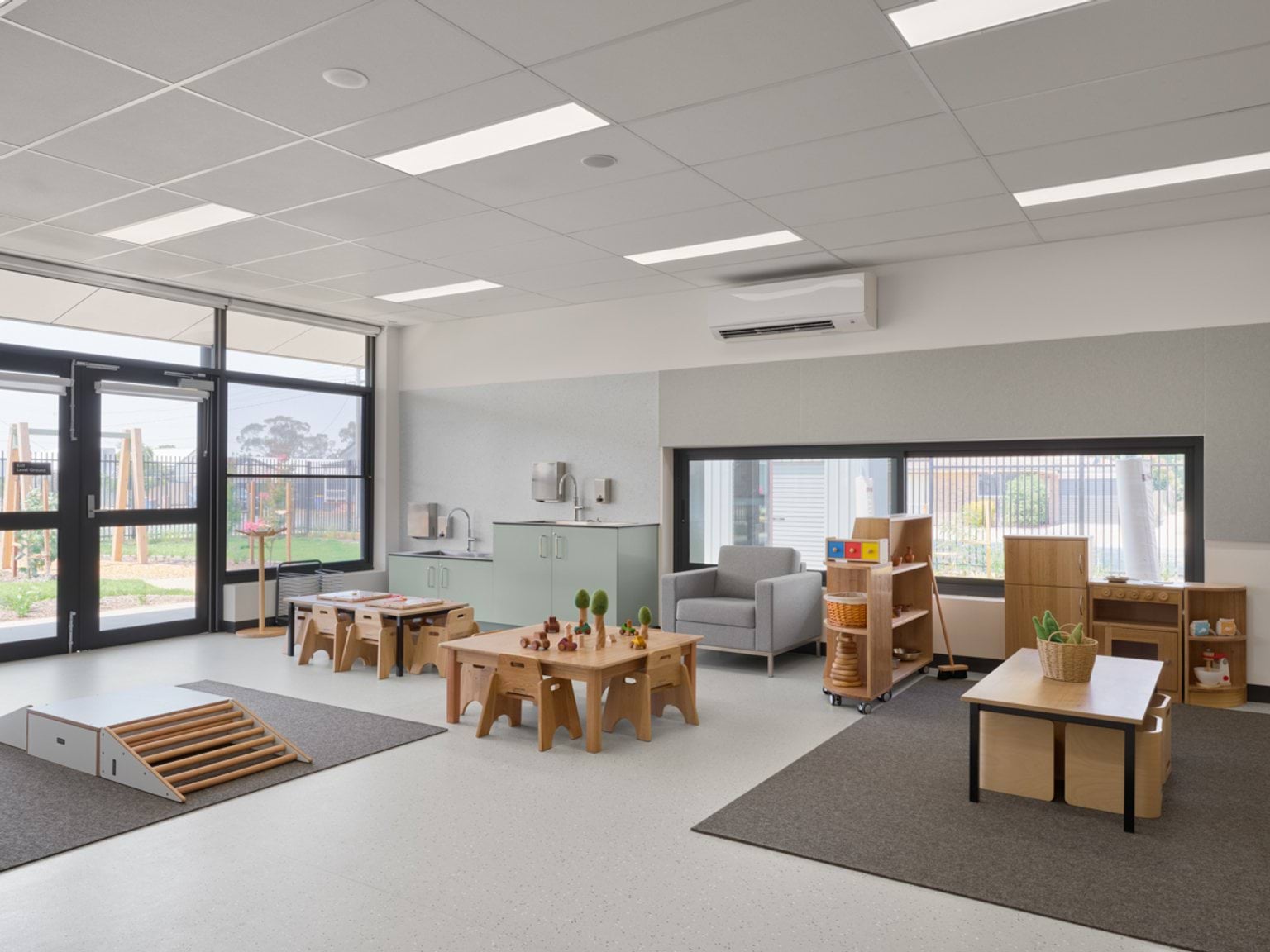 An early learning room with small tables, soft play mats, a reading corner, and views of the outdoor play area.