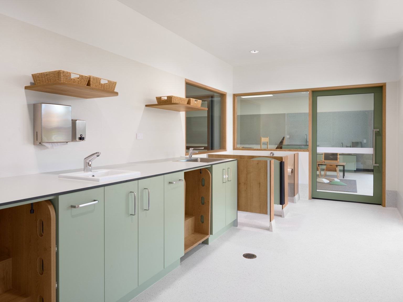 A staff preparation room with sinks, green cabinetry, open shelving, and a window into an adjoining learning space.