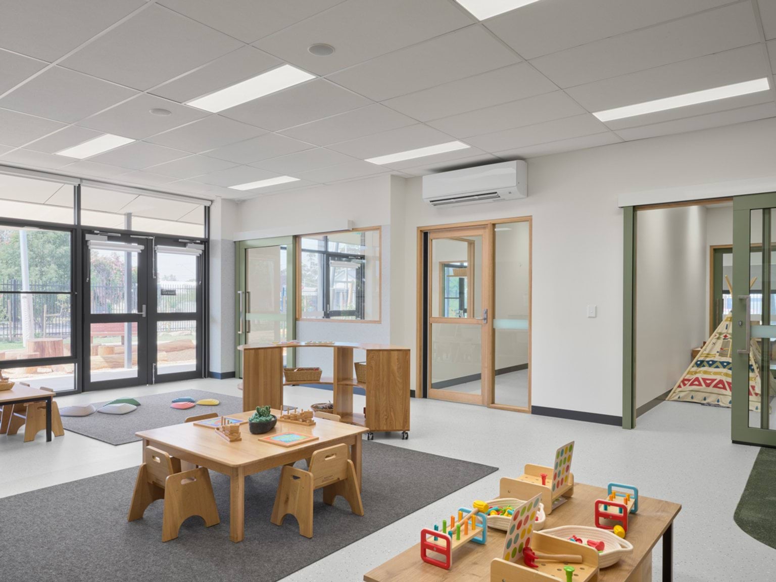 A spacious early learning room with child‑sized tables, timber play furniture, and views to the outdoor play area.