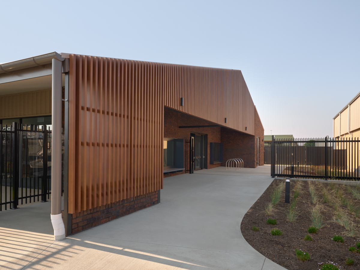 An exterior view of an early learning centre entrance with timber‑screen cladding, paved pathways, and landscaped planting.