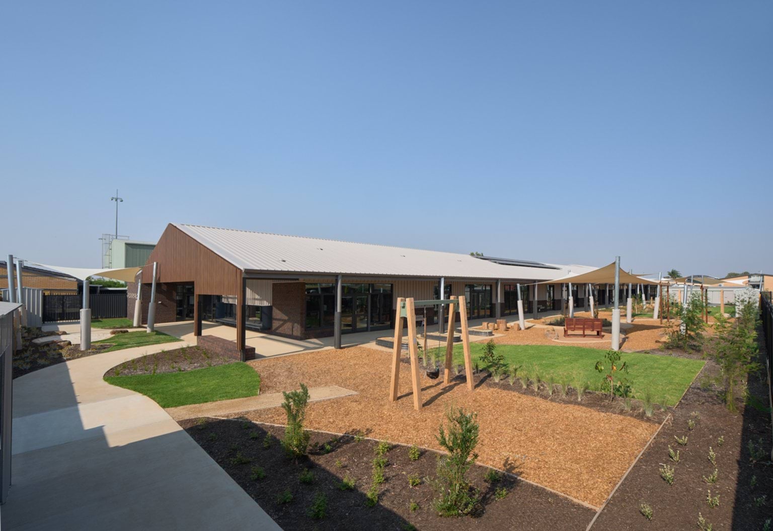 A wide view of the early learning centre showing outdoor play spaces, lawn areas, shade sails, and adjoining single‑storey buildings.