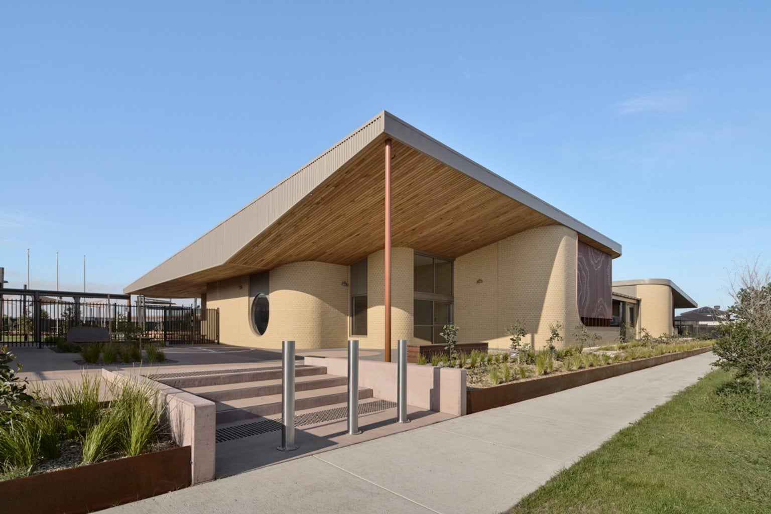 An exterior view of the early learning centre showing a curved brick facade, timber soffit, landscaped garden beds, and an entry path.